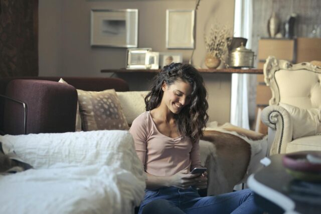 Photo of Smiling Woman Sitting on the Floor While Using Her Phone