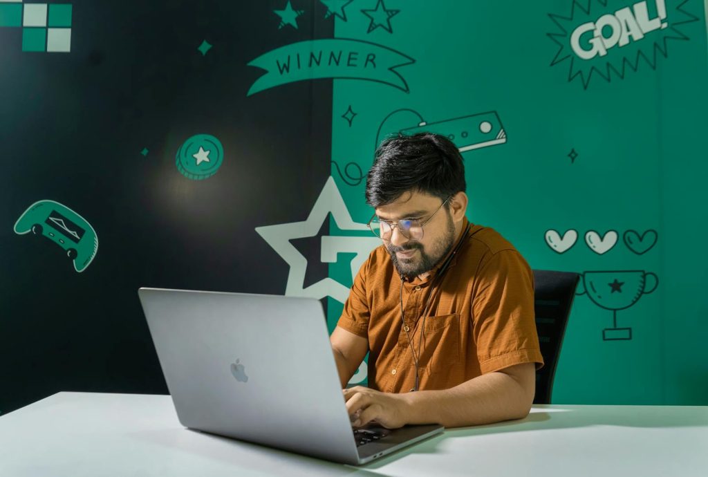 A man with a beard and glasses smiles while gaming on a laptop indoors, decorated with gaming graphics.